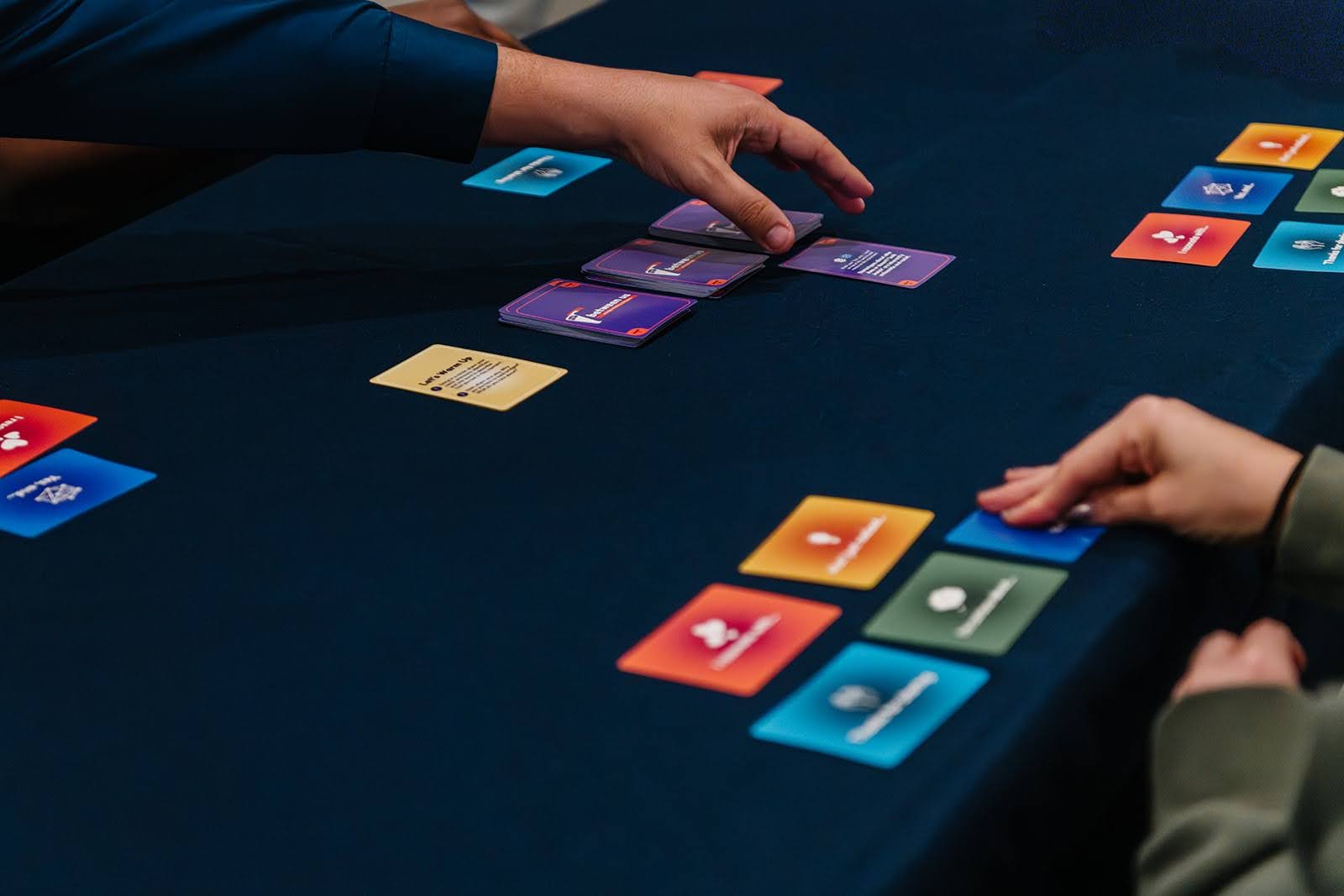 Two people’s hands over a table with Between Us set up, of 3 piles of prompt cards, and 5 colorful impact cards